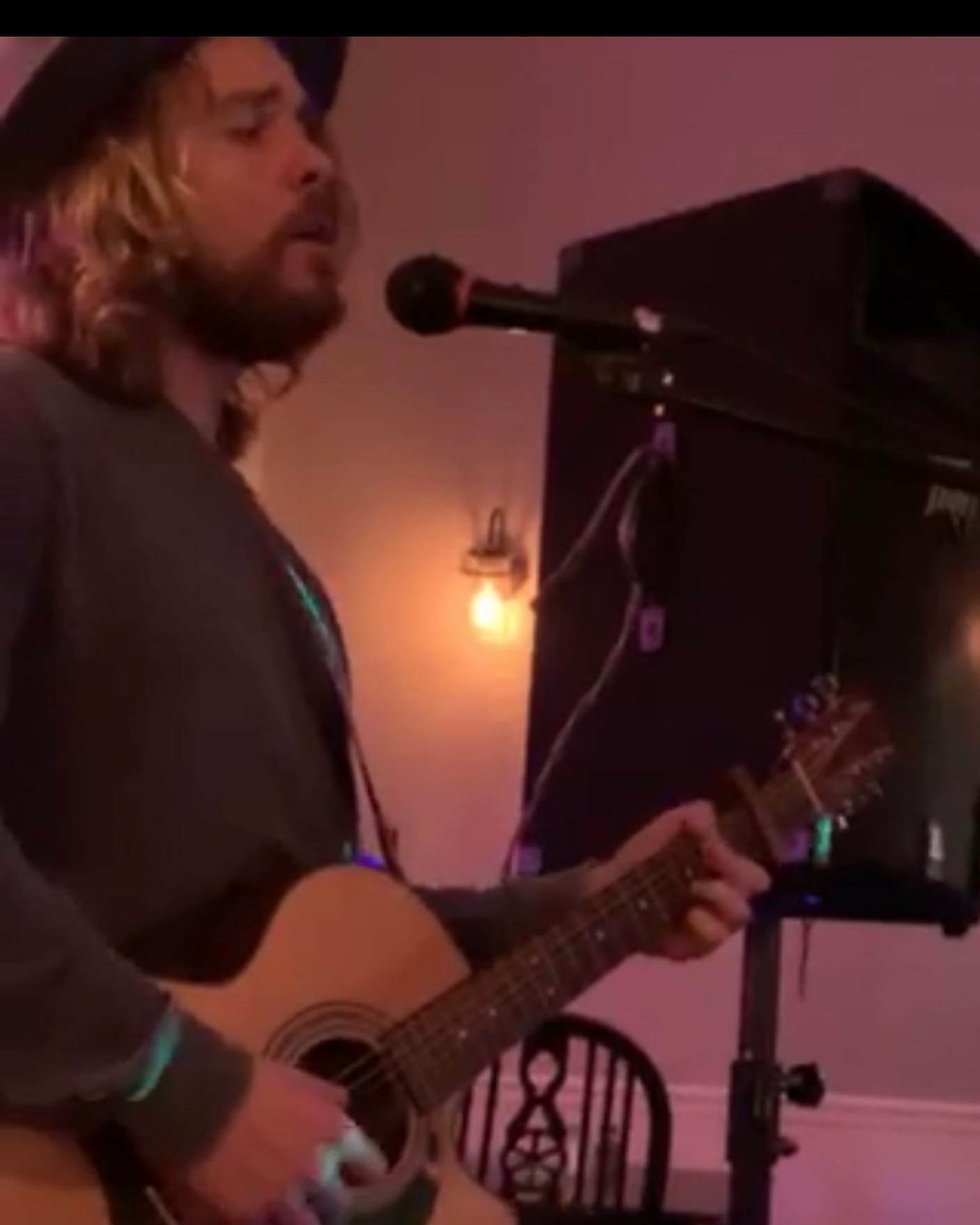 A man with long hair singing into a microphone while playing an acoustic guitar in a dimly lit room.