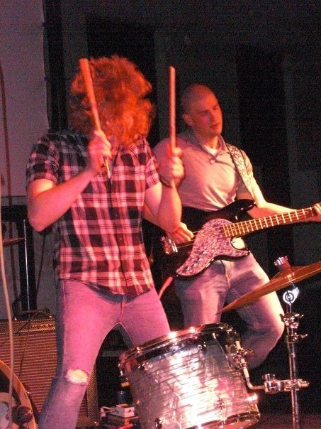 Two musicians performing on stage, one playing guitar and one on drums.
