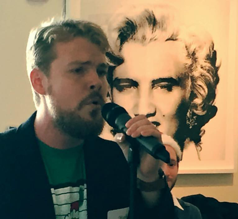 A close-up of a man's face singing into a microphone next to an illustrated or painted portrait.