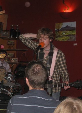 A person with shaggy hair wearing a plaid shirt singing into a microphone on stage, with audience members visible in the foreground.